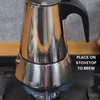 Polished metal Moka Pot with a black handle on a gas stove flame. Text: Place on stovetop to brew.