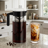 Tall glass Cold Brew Maker with black lid and a glass of iced coffee with milk on a granite kitchen counter.